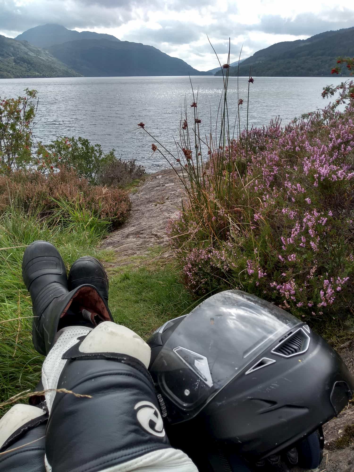 Legs of author wearing bike trouser leathers and boots, with helmet next to boots, surrounded by heather and looking out over Loch Lomond