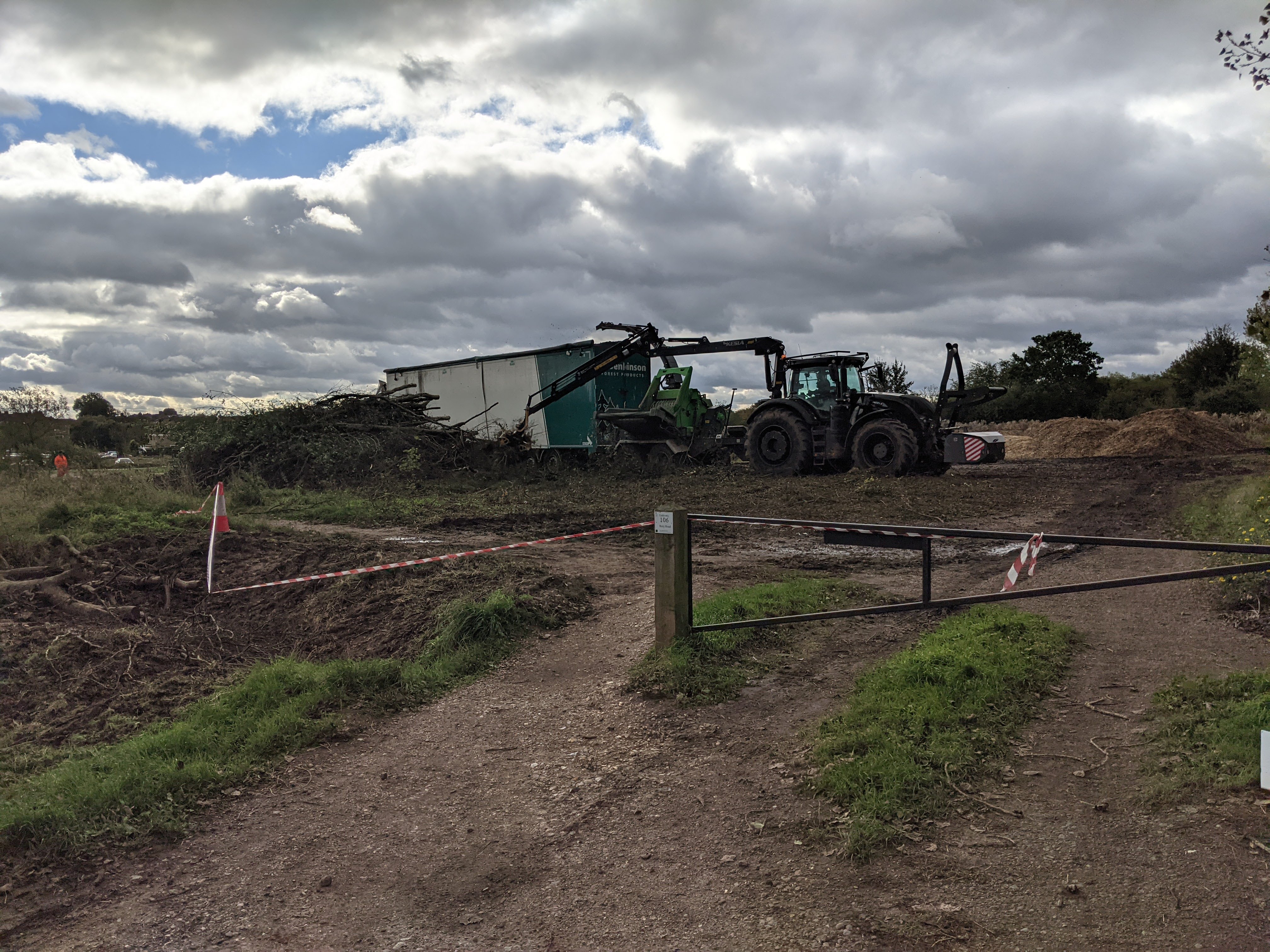 Pile of trees on the ground next to a tractor, which seems to be lifting them into a device which is shredding them, into a lorry