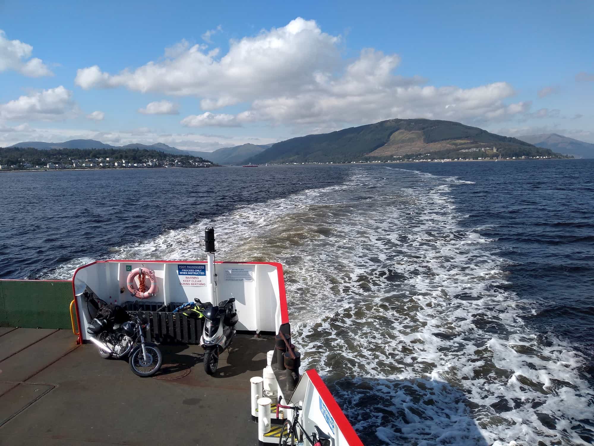 My motorbike on the back of a ferry, with coastal hills in the distance and lots of water turbulence behind the ferry
